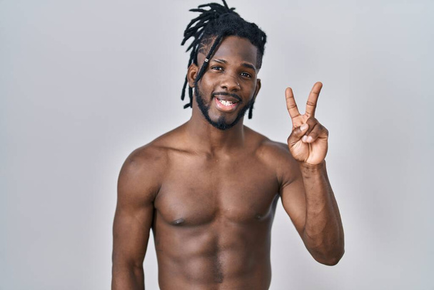 Afričan s dredy, stojící bez košile nad izolovaným pozadím a usmívající se s veselou tváří, mrkající na kameru a předvádějící znamení vítězství. číslo dvě.  - Fotografie, Obrázek