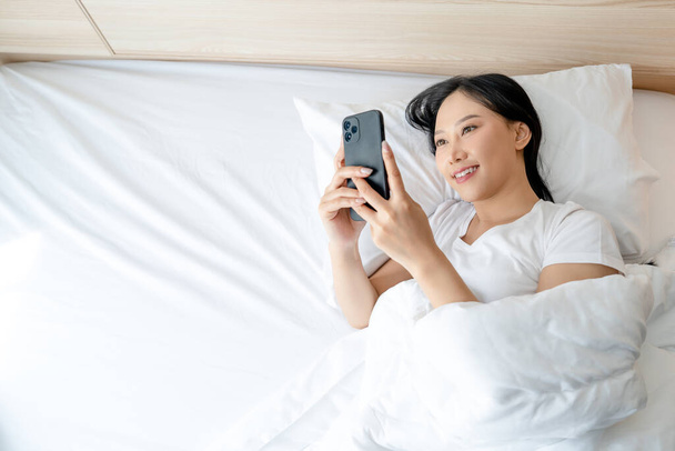 Mulher asiática bonita usando smartphone na cama branca de manhã com espaço de cópia. Conceito de dependência móvel. - Foto, Imagem