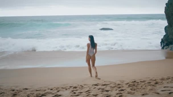 Sexy jong meisje stappen op het strand zand dragen van witte badpak achteraanzicht. Onbekende aantrekkelijke vrouw loopt naar schuimige oceaan golven bewolkte dag. zorgeloze ontspannen dame genieten van mooie zeegezicht sombere dag. - Video