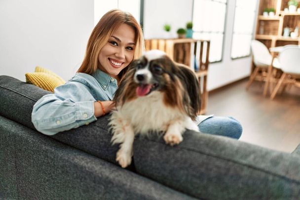 Fiatal fehér nő mosolygós magabiztos ül kanapén kutyával otthon - Fotó, kép