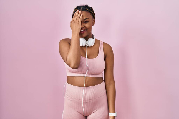 African american woman with braids wearing sportswear and headphones covering one eye with hand, confident smile on face and surprise emotion.  - Photo, image