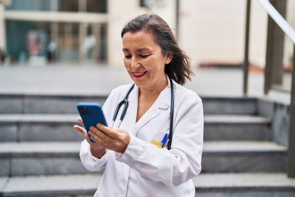 Middle age woman wearing doctor uniform using smartphone at street - Zdjęcie, obraz