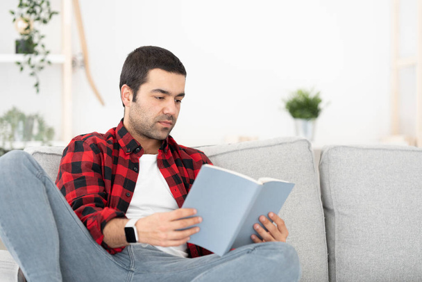 Ισπανόφωνος νεαρός κάθεται στον καναπέ και διαβάζει ένα βιβλίο. Μαθητεία σπουδαστών στο σπίτι. - Φωτογραφία, εικόνα