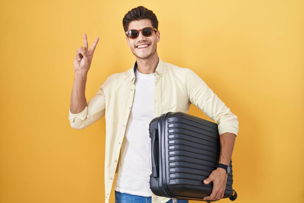 Junger hispanischer Mann mit Koffer, der in den Sommerurlaub geht, lächelt in die Kamera und zeigt Finger, die Siegeszeichen machen. Nummer zwei.  - Foto, Bild