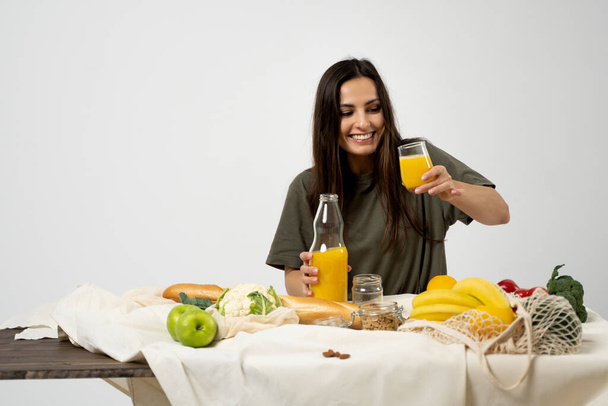 Woman in green t-shirt pouring a juice from a glass bottle in a glass over a table with mesh eco bag, healthy vegan vegetables, fruits, bread, snacks. Zero waste concept - 写真・画像