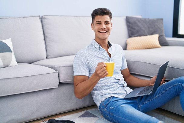 Young hispanic man using laptop and drinking coffee sitting on floor at home - Foto, imagen