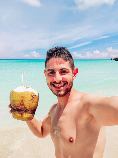 Cheerful shirtless guy holding coconut with drinking straw near serene sea and taking selfie during trip in Cuba in sunshine - Foto, immagini