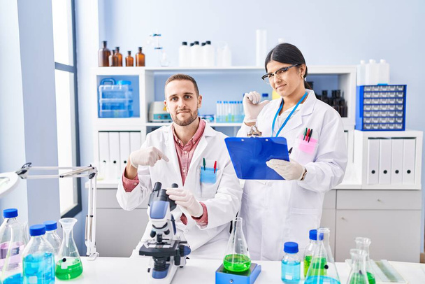 Двое молодых людей, работающих в научной лаборатории, указывая пальцем на одного, улыбающегося счастливого и гордого  - Фото, изображение