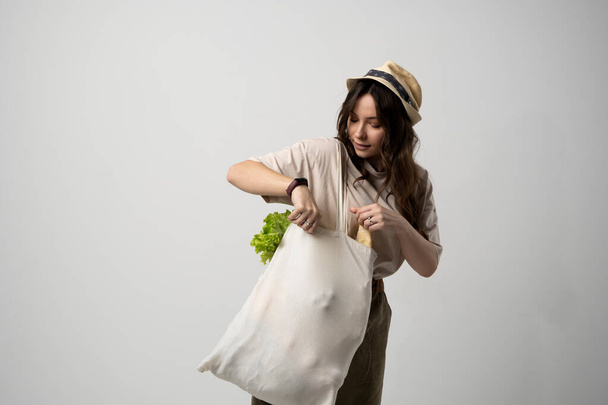 Smiling woman in a t-shirt and a hat holding reusable cotton shopping bag with vegetables, bread and greens. Concept of no plastic, zero waste, plastic free, sustainable lifestyle - Fotó, kép