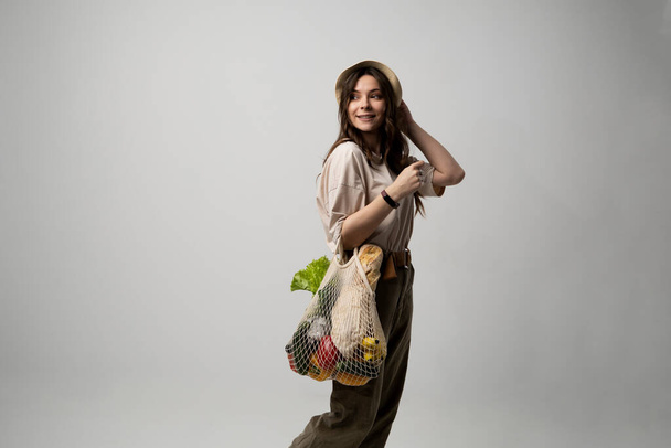 Concept of no plastic. Zero waste, plastic free. Sustainable lifestyle. Happy woman in a beige t-shirt and a hat holding reusable cotton shopping bag with organic groceries, bread and greens - Foto, imagen