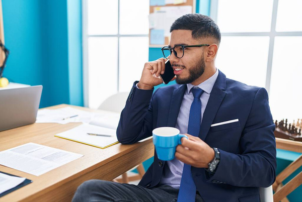Молодой латинский бизнесмен разговаривает по смартфону, пьет кофе в офисе - Фото, изображение