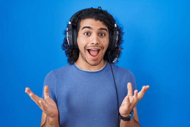 Hispanic man with curly hair listening to music using headphones celebrating crazy and amazed for success with arms raised and open eyes screaming excited. winner concept  - Photo, image