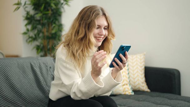 Mujer rubia joven usando teléfono inteligente sentado en el sofá en casa - Foto, Imagen