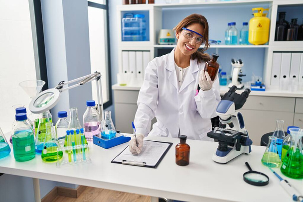 Νεαρή Λατίνα γυναίκα που φοράει στολή επιστήμονα και γράφει στο πρόχειρο στο εργαστήριο - Φωτογραφία, εικόνα