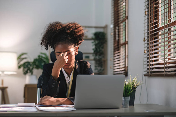 Joven mujer afroamericana con peinado afro se ve molesta y estresada, sentada en el escritorio, usando una computadora portátil, pensando y mirando a la cámara, sintiéndose cansada y aburrida con problemas de depresión.. - Foto, Imagen