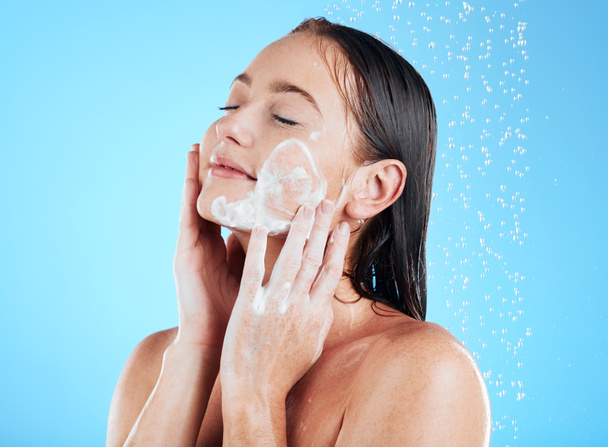 Ντους, πρόσωπο και γυναίκα πλύσιμο προσώπου με σαπούνι σε μπλε φόντο για περιποίηση, καθαρισμό και ευεξία. Περιποίηση δέρματος, μπάνιο και χαρούμενη κοπέλα με αφρό για υγιεινή, ενυδάτωση και ομορφιά στο studio. - Φωτογραφία, εικόνα