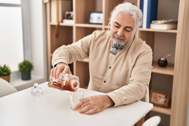 Middle age grey-haired man pouring whisky on glass sitting on table at home - Fotoğraf, Görsel