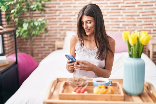 Mujer hispana hermosa joven desayunando con un smartphone en el dormitorio - Foto, Imagen