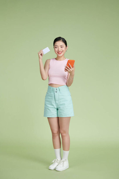 Retrato de una mujer sonriente con tarjeta de crédito y teléfono móvil sobre verde - Foto, imagen