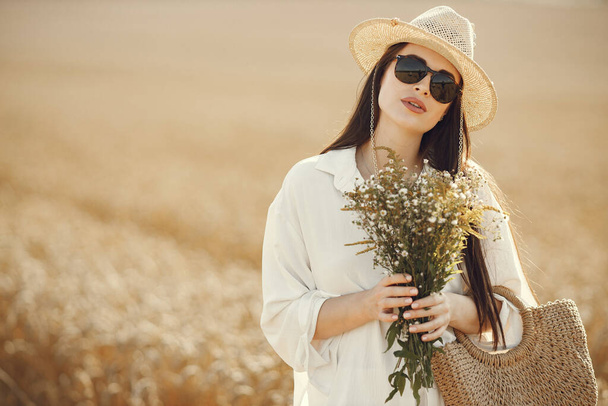Pregnant woman holding wildflowers bouquet in straw bag, walking in wheat field. Brunette woman walking in summer field wearing white clothes. Woman wering sunglasses, straw hat and white costime. - Фото, зображення