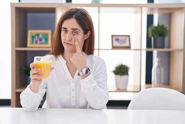 Mujer morena bebiendo un vaso de jugo de naranja apuntando al ojo observándote gesto, expresión sospechosa  - Foto, Imagen