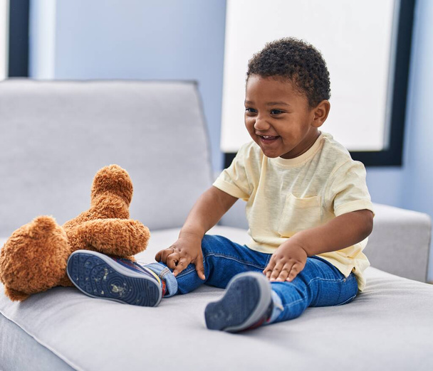 Niño afroamericano jugando con oso de peluche sentado en el sofá en casa - Foto, imagen