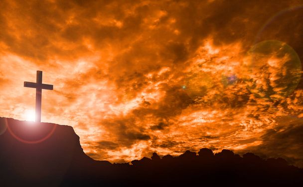 Krzyż na pomarańczowym niebie z chmurami i promieniami słońca nad urwiskiem - Zdjęcie, obraz
