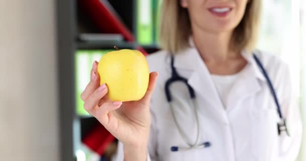 La doctora positiva sostiene manzana fresca en la mano sonriendo en la clínica. Mujer rubia alegre en uniforme médico recomienda una dieta saludable - Imágenes, Vídeo