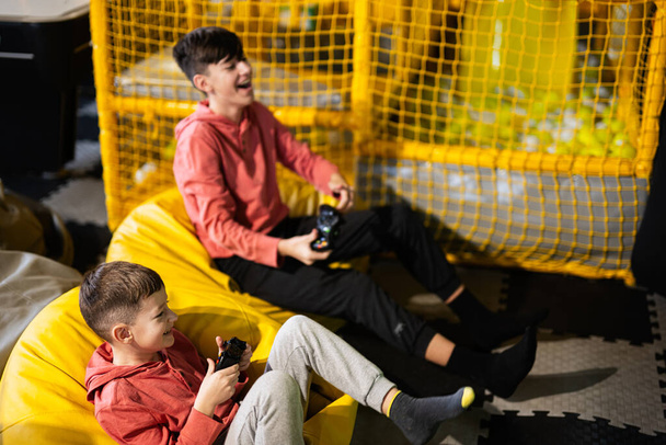 Dos hermanos jugando videoconsola, sentados en puf amarillo en el centro de juego de los niños. - Foto, imagen