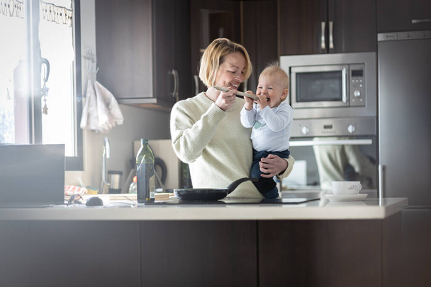 Gelukkige moeder en kleine baby jongen koken en proeven van gezond eten in de huiselijke keuken. Familie, levensstijl, huishoudelijk leven, voedsel, gezond eten en mensen concept - Foto, afbeelding
