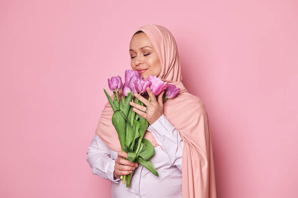 Charmante junge schwangere Muslimin aus dem Nahen Osten im rosafarbenen Hijab, schnüffelt an einem Strauß Tulpen und posiert mit geschlossenen Augen vor isoliertem rosafarbenem Hintergrund. Konzept zum Internationalen Frauentag und Muttertag - Foto, Bild
