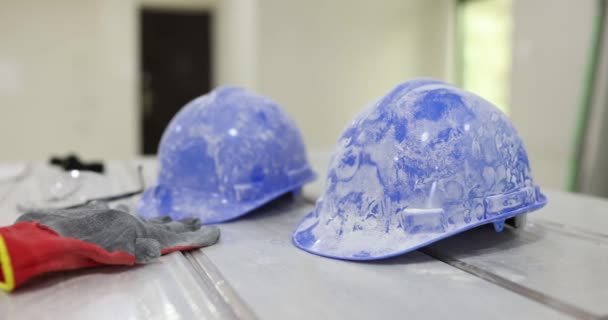 Bouwers hardhoeden en handschoenen bedekt met wit stof op tafel in gerestaureerde kamer. Veiligheidsvoorzieningen op de bouwplaats. Blauwe plastic vuile helmen - Video