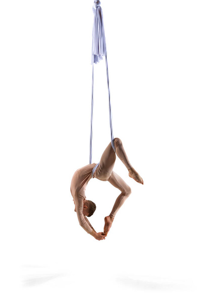 Grace and flexibility. Young man, professional aerial gymnast athlete making performance on silk aerial ribbons against white studio background. Concept of art, sportive lifestyle, action and motion - Fotó, kép