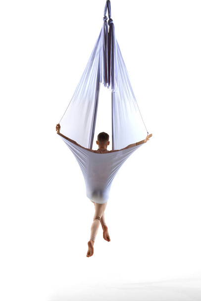 Professional male aerial gymnast training with transparent silk fabric against white studio background. Young sportsman. Concept of art, sportive lifestyle, hobby, action and motion, beauty. Back view - Fotografie, Obrázek
