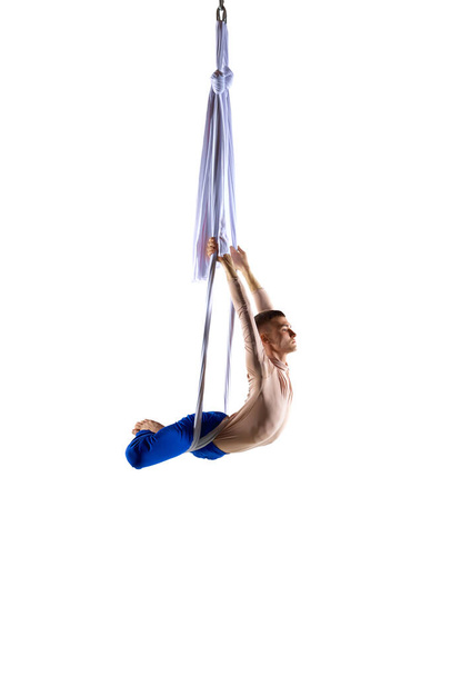 Strong, athletic young man, professional aerial gymnast, acrobat training with aerial tissues against white studio background. Concept of art, sportive lifestyle, hobby, action and motion, beauty - Фото, изображение