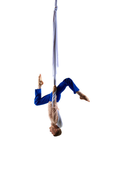 Upside down. Young male professional aerial gymnast, acrobat training on aerial fabric against white studio background. Concept of art, sportive lifestyle, hobby, action and motion, beauty - 写真・画像
