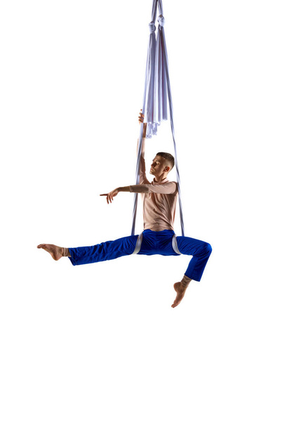 Artistic performance. Young man, professional aerial acrobat doing gymnastics tricks on aerial tissue against white studio background. Concept of art, sportive lifestyle, hobby, action, motion, beauty - Foto, imagen