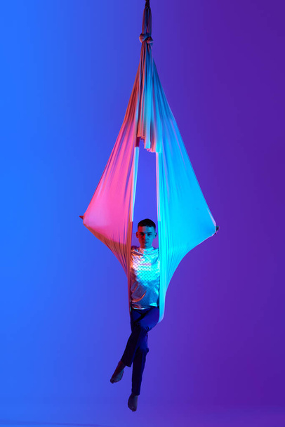 Athletic young man, professional aerial gymnast doing tricks with ribbons against gradient blue purple background in neon light. Butterfly. Concept of art, sportive lifestyle, hobby, action and motion - Valokuva, kuva