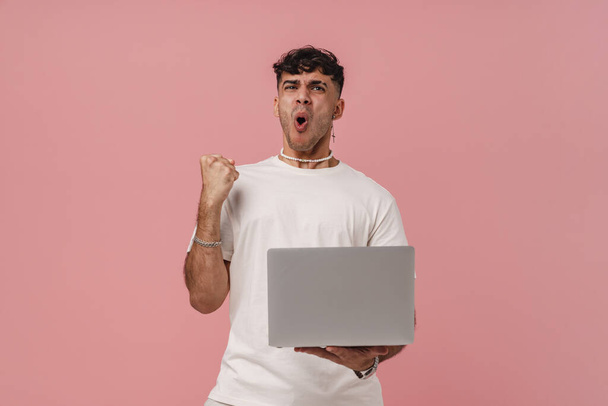 Young handsome stylish enthusiastic man holding laptop doing winner gesture with raised fist and opened mouth , while standing over isolated pink background - Foto, immagini