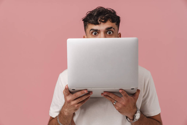 Young handsome curly man peeking from behind a laptop and looking at camera, while standing over isolated pink background - Photo, image
