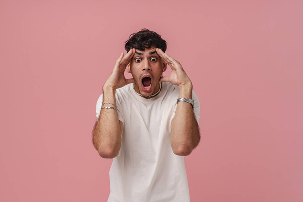 Portrait of young handsome stylish shocked man with wide opened eyes and mouth, touching his head both hands and looking downward , while standing over isolated pink background - Fotografie, Obrázek