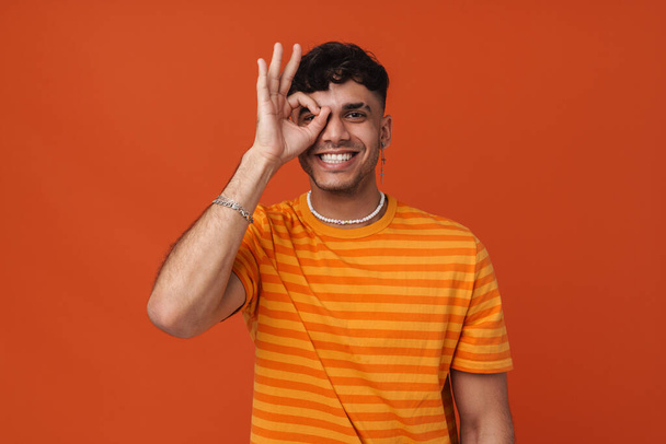 Young handsome stylish happy smiling man doing ok gesture on his eye and looking at camera ,while standing over isolated orange background - Φωτογραφία, εικόνα