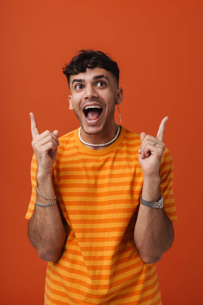 Young handsome happy enthusiastic man poiting upwards both hands and looking aside, while standing over isolated orange background - Фото, зображення
