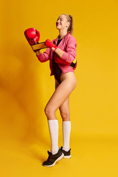 Sporty girl. One beautiful girl putting on boxing gloves with pleasure facial expression over yellow background. Concept of sport, hobby, fitness, fashion, human emotions, beauty - Foto, imagen