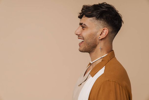 Brunette young man with earring laughing and looking aside isolated over beige background - Foto, Imagen