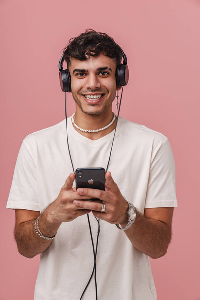 Brunette young man in headphones smiling while using mobile phone isolated over pink background - Foto, afbeelding