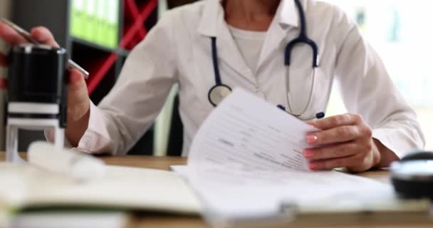 Vrouwelijke dokter nietjes document papieren zitten aan tafel in het ziekenhuis kantoor. Vrouw met stethoscoop werkt met patiënt geschiedenis record in kliniek slow motion - Video