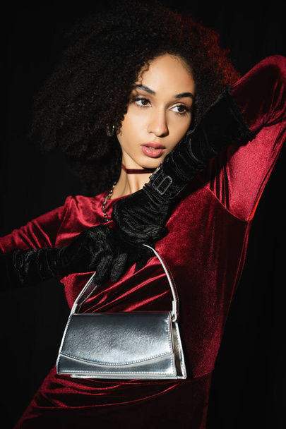 Trendiges afrikanisch-amerikanisches Model in Samtkleid und Handschuhen, die silberne Handtasche isoliert auf schwarz halten  - Foto, Bild