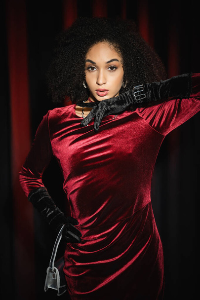 Pretty african american model posing in velvet dress and gloves near burgundy drapery  - Foto, afbeelding