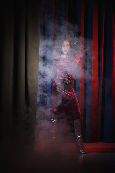 Elegantní africká americká žena v šatech a rukavicích na červeném pozadí s modrým světlem a kouřem  - Fotografie, Obrázek